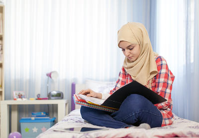 Young student in head wrap reading materials in their lap seated on a bed.
