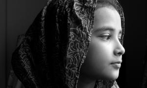In a black and white photograph, a young girl wearing a head covering looks pensively off camera 