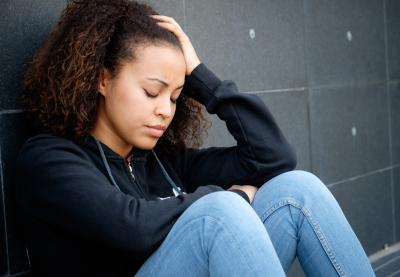 Young person of color in visible distress sitting with their back against a wall, hand on their head.