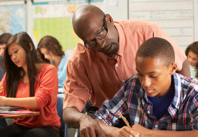 Teacher of color guiding student of color over their shoulder in a classroom setting.