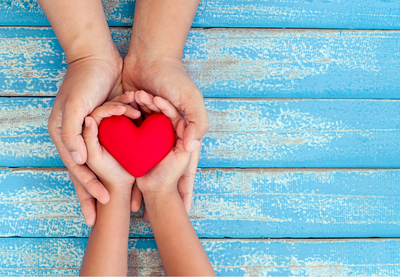 A pair of larger hands encircles a child's hands and in the child's hands is a red heart.