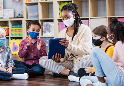 A teacher and a group of young students, all wearing masks, gather around in a half circle so they can look at images on an electronic tablet and listen during what appears to be story time. 