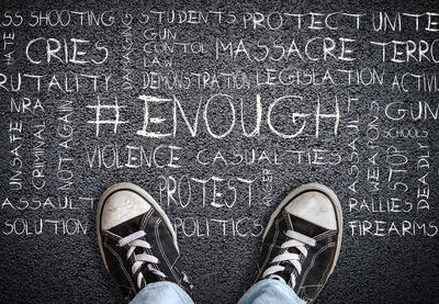 A child's feet with sneakers on them stand in front of words that include protect, students, mass shooting, cries, brutality, activism, walkout and more etched in chalk on the ground. 