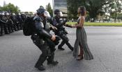 A female BlackLivesMatter protester in a long dress looks straight ahead as two police officers in riot gear approach her. 